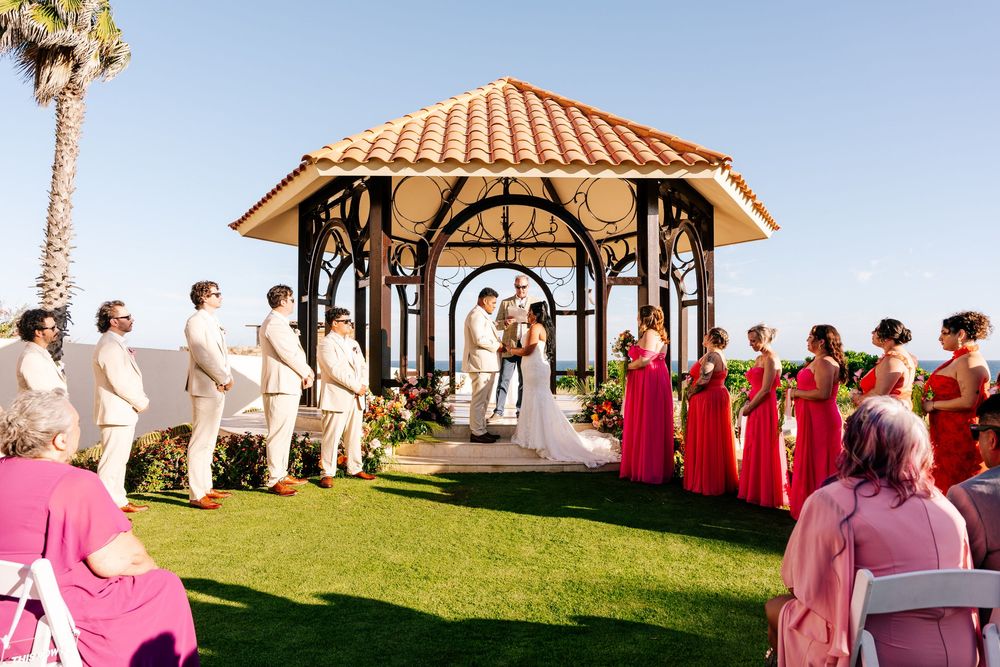 Outdoor beachside wedding ceremony under a tiled-roof gazebo with groomsmen in white and bridesmaids in coral dresses on a green lawn.