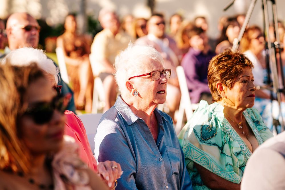 Elderly woman with white hair and red glasses sits attentively among a crowd of seated guests at an outdoor event.