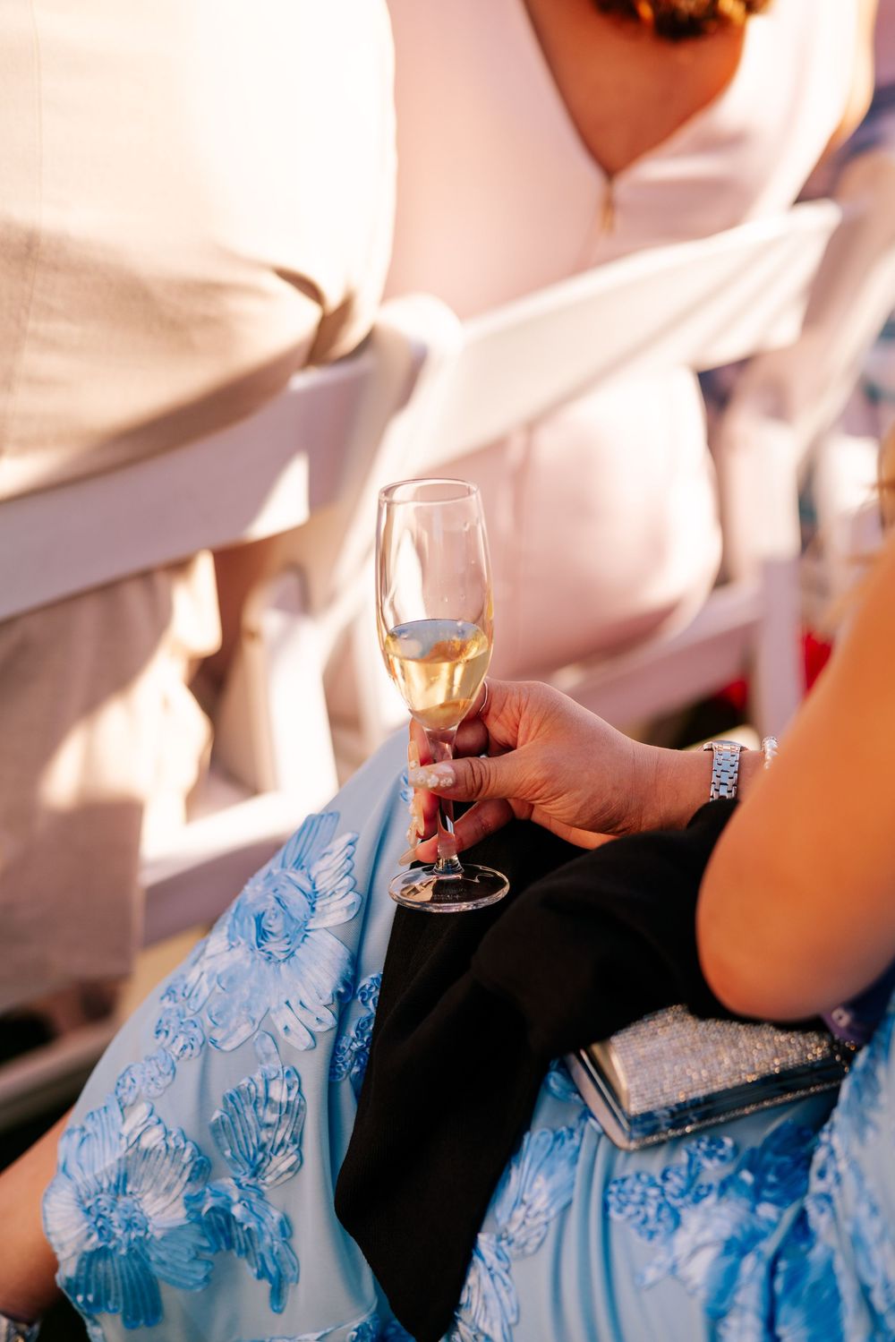 Woman in a blue floral lace dress holds a glass of champagne beside a sparkly clutch while seated at an outdoor wedding ceremony.