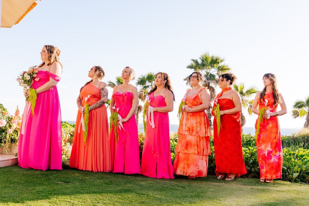 Seven bridesmaids in vibrant pink, orange, and coral dresses hold bouquets while standing on a green lawn with palm trees behind them.