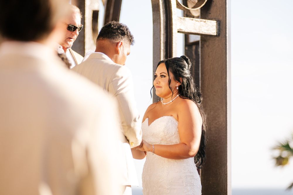 A bride in a white strapless wedding gown holds hands with her groom during an outdoor beachside ceremony as an officiant looks on.