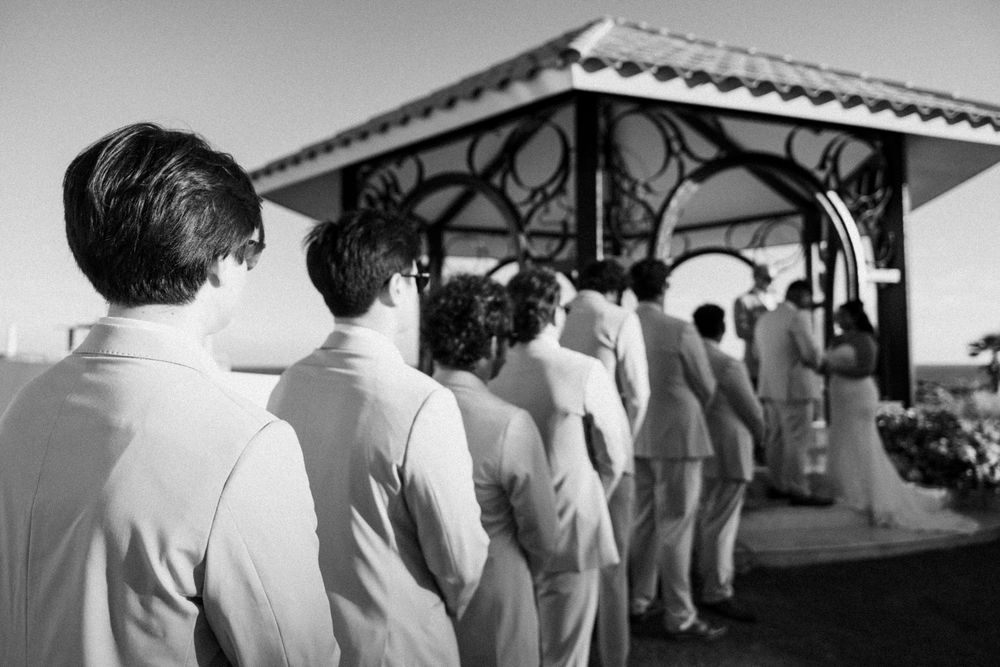 Groomsmen in light suits stand in a row facing a gazebo during a beachside wedding ceremony, in black and white.
