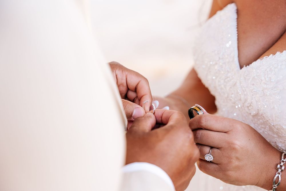 Bride in a beaded white gown exchanges wedding rings with the groom during a close-up ceremony moment, her diamond engagement ring visible.