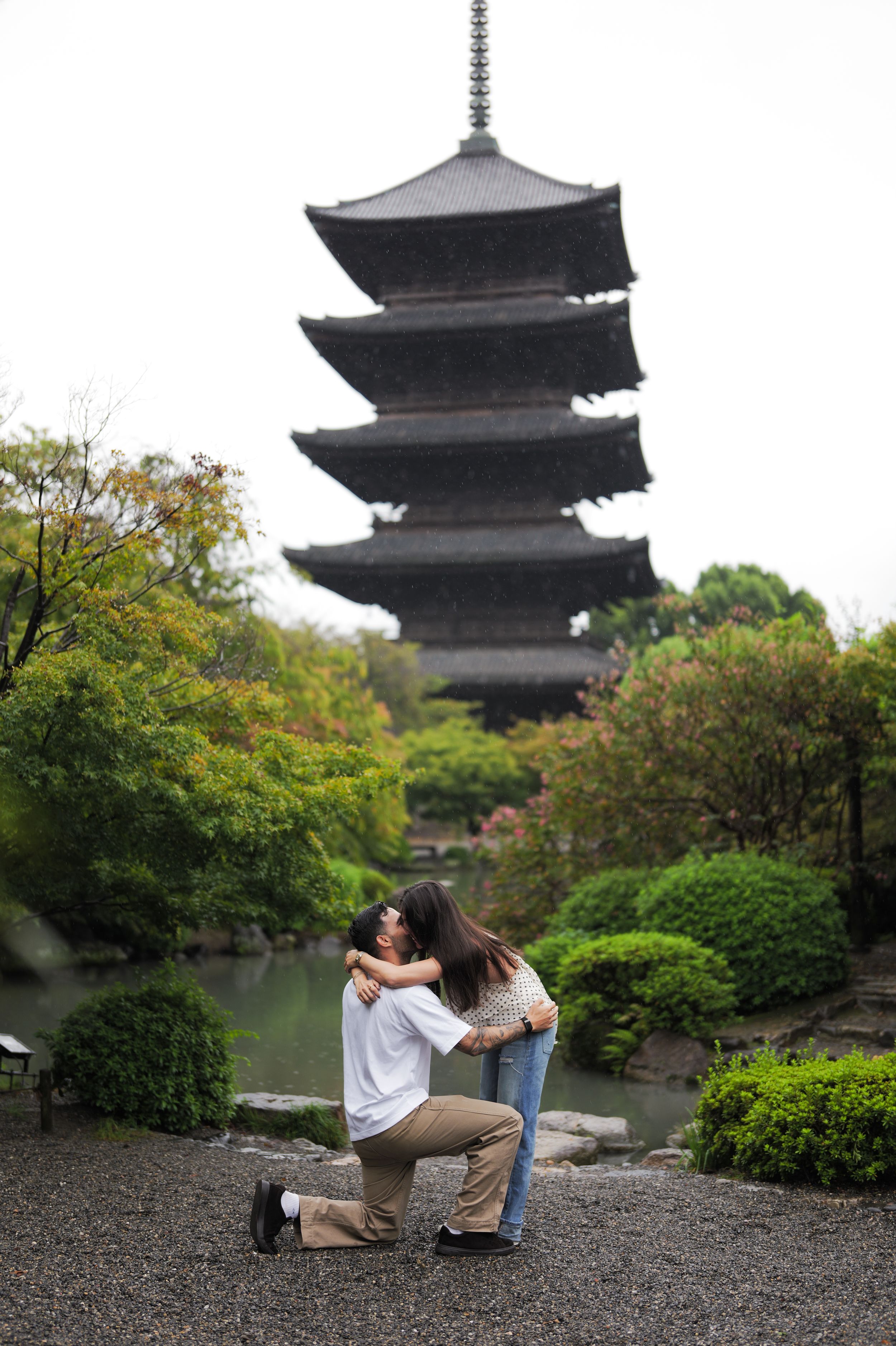 Couple embracing and kissing as one kneels in a marriage proposal beside a pond, with a tall Japanese pagoda and green trees behind them.
