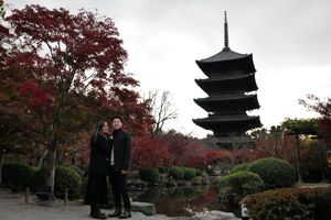 Couple in dark coats pose beside a pond near vibrant red autumn maples, with a tall five-story Japanese pagoda rising under an overcast sky.