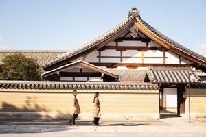 Two people in long brown coats walk past a traditional Japanese temple with a tiled roof and ochre wall under a clear blue sky.