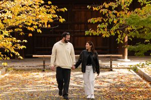 Smiling couple holding hands walking along a leaf-covered path under golden autumn trees in a peaceful park.