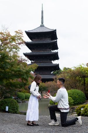Man kneeling on one knee proposes to a woman in a white dress before a tall five-story pagoda surrounded by autumn trees.