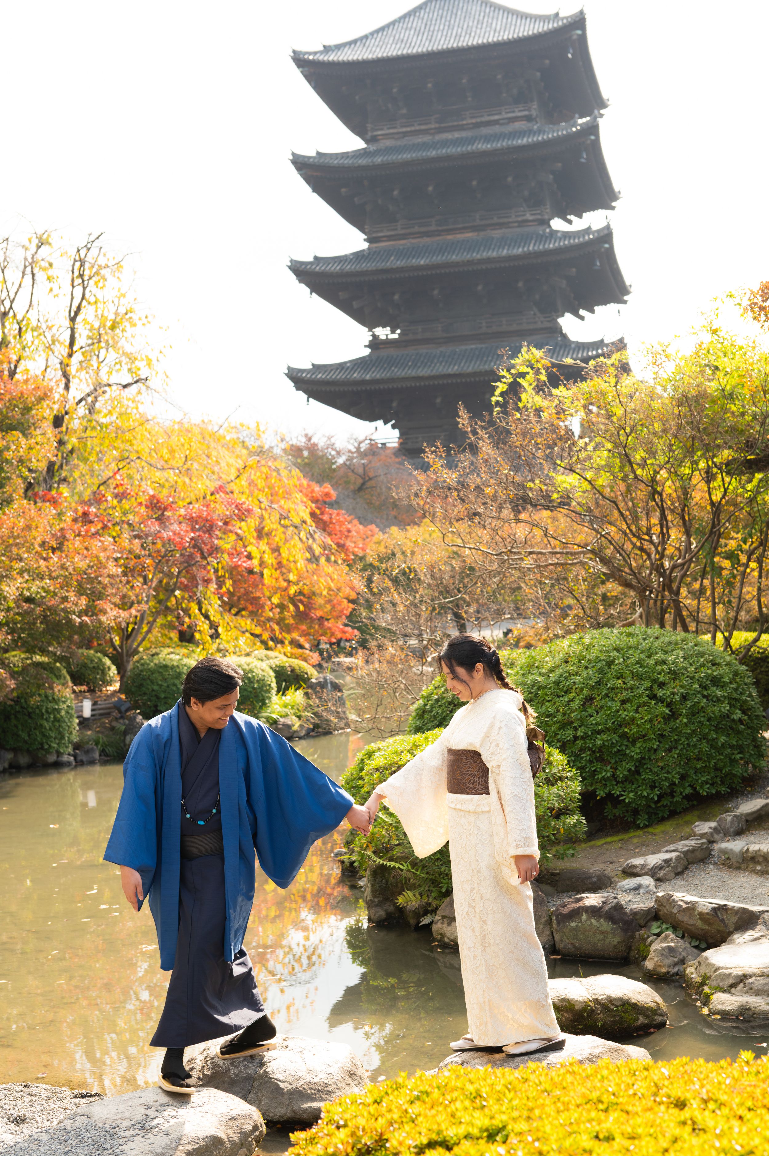 Couple in traditional Japanese kimono holding hands by a pond with autumn foliage and a historic multi-tiered pagoda in the background.