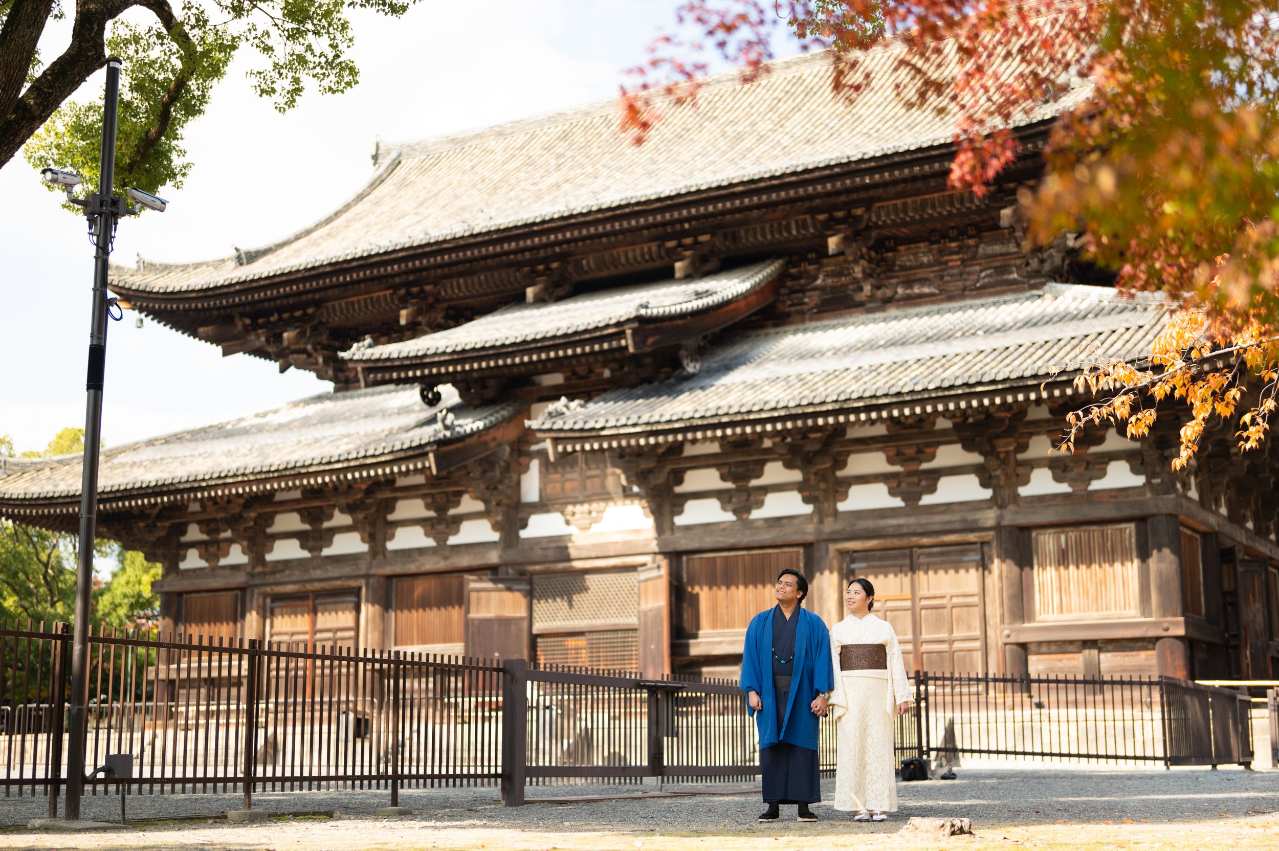 Couple in traditional Japanese kimono clothing standing before a historic wooden temple surrounded by colorful autumn foliage.