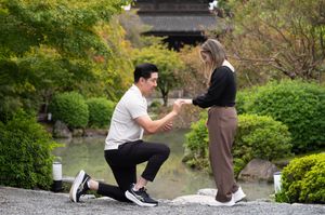 Man kneeling on one knee proposing to a smiling woman beside a peaceful pond in a lush Japanese garden with a pagoda in the background.