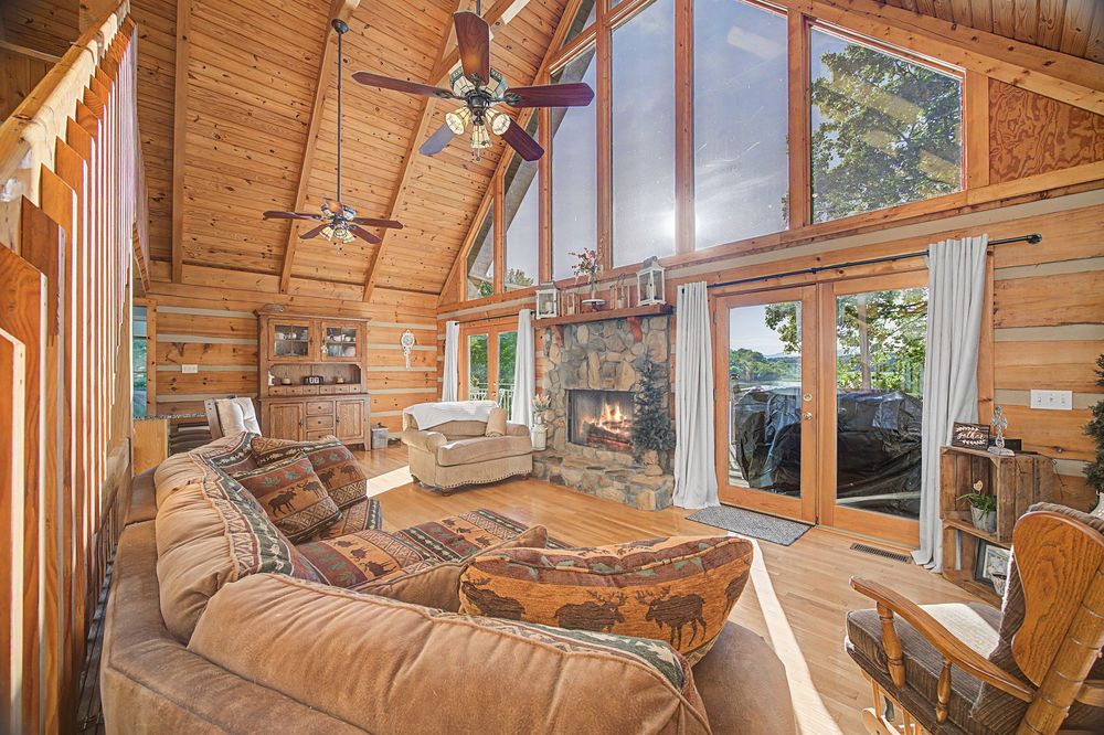 Cozy log cabin living room with vaulted wood ceiling, stone fireplace, leather sofa, and large windows offering scenic views.