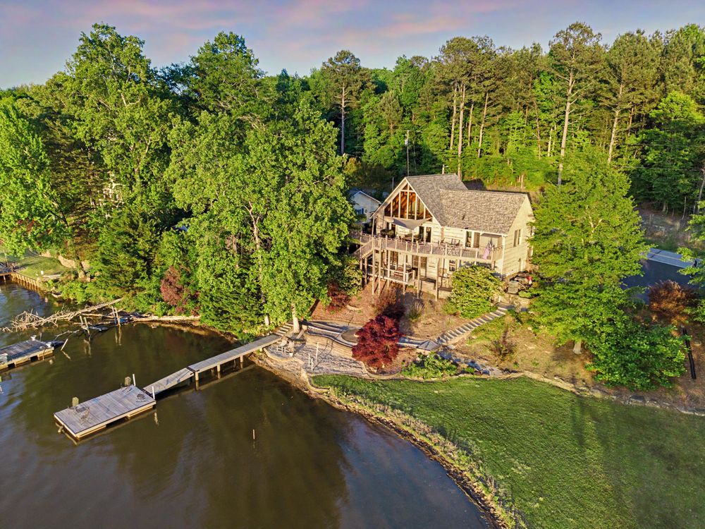 Aerial view of a lakefront home surrounded by lush green trees with a private dock on calm waters.