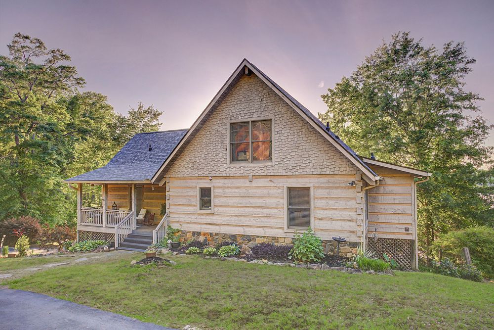 Rustic log cabin home surrounded by lush green trees with a covered porch and manicured landscaping at dusk.
