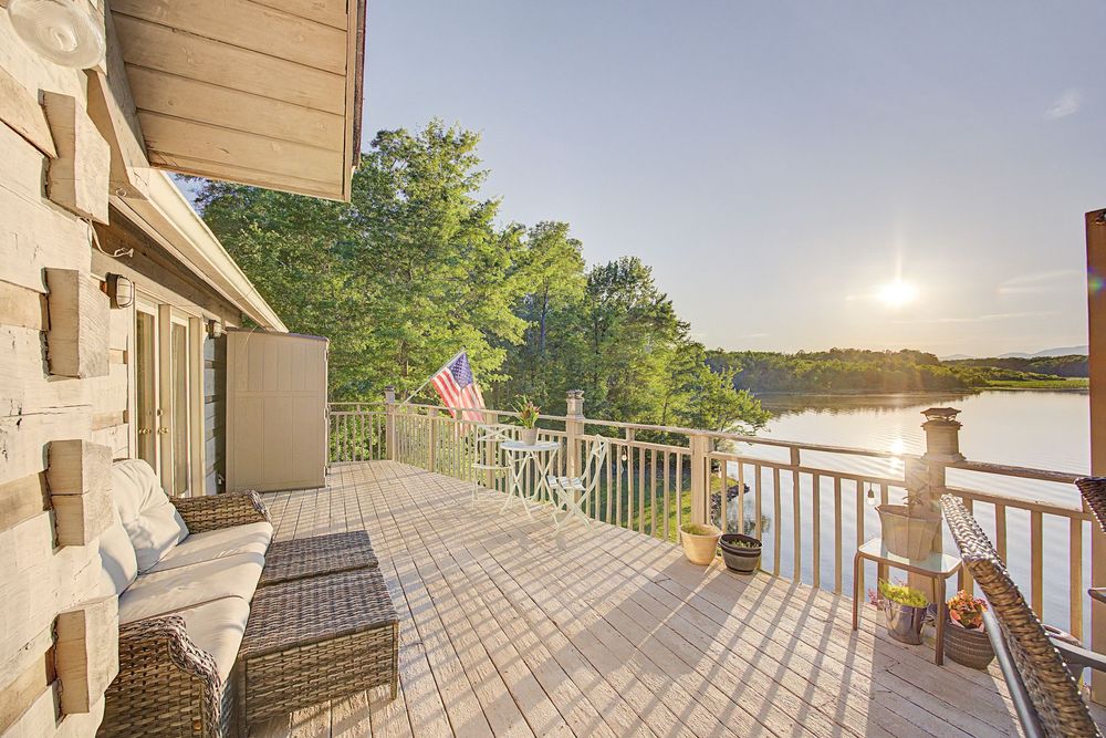 Spacious wooden deck overlooking a serene lake at sunset, featuring wicker furniture and an American flag.