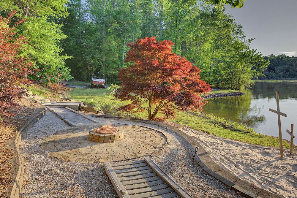 Scenic lakeside landscape with curved stone path, red Japanese maple tree, fire pit, and lush green forest in background.