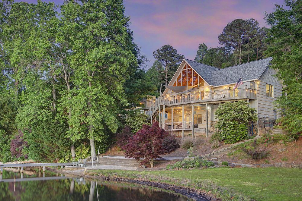 Charming lakeside cabin with large deck surrounded by lush trees at dusk, reflecting warm lights on calm water.