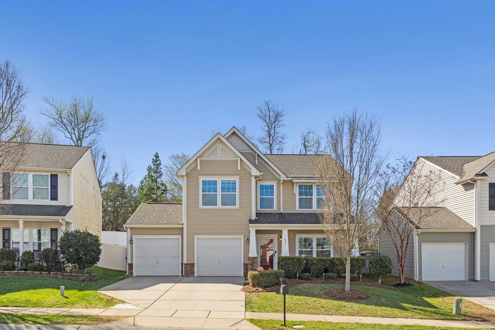 Two-story beige suburban home with double garage, manicured lawn, and mature trees on a sunny day.