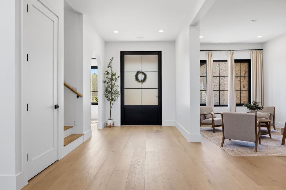 Bright modern entryway with light hardwood floors, black glass-panel front door, white walls, and open dining area in background.