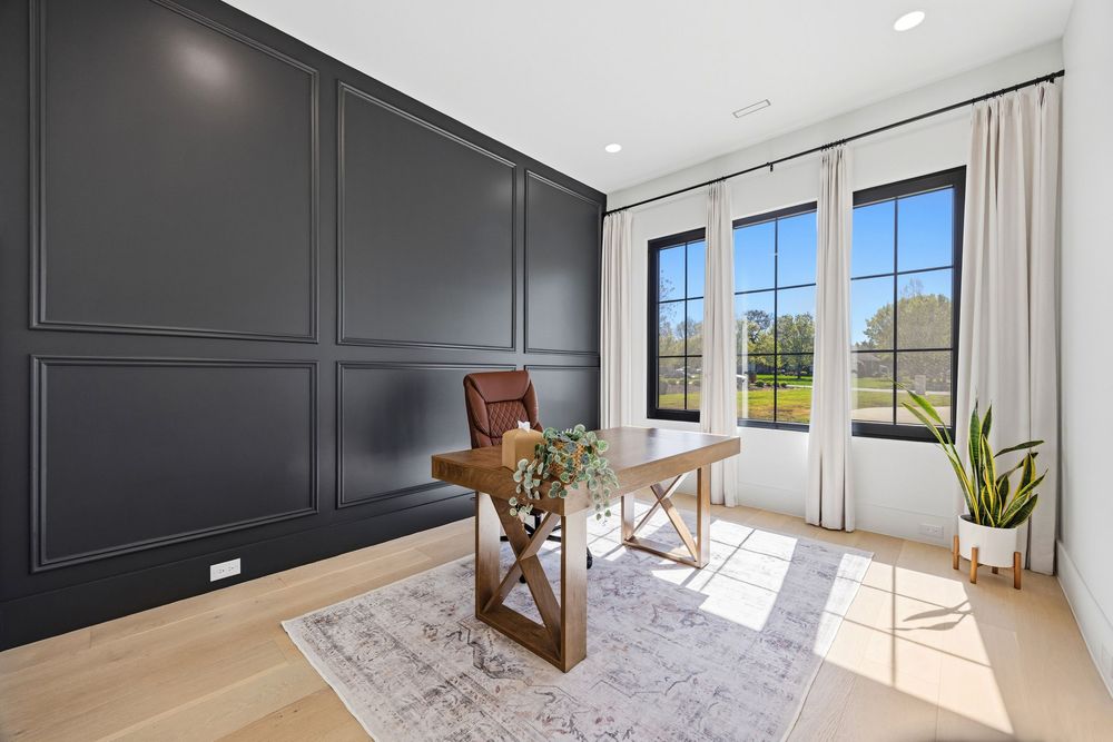 Modern home office with dark paneled accent wall, wooden desk, leather chair, and large windows overlooking greenery.