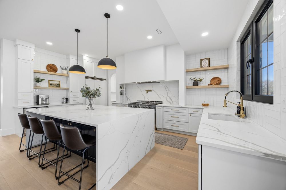 Modern white kitchen with marble island, pendant lights, bar stools, open shelving, and hardwood floors.