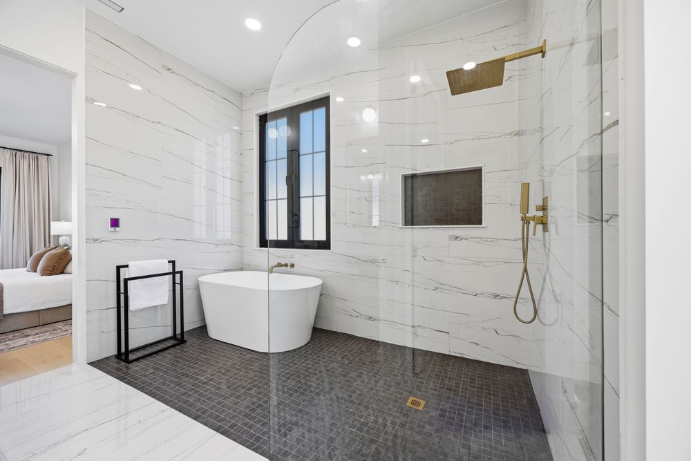 Luxury bathroom with freestanding soaking tub, gold fixtures, glass shower, and white marble walls with dark mosaic tile floors.