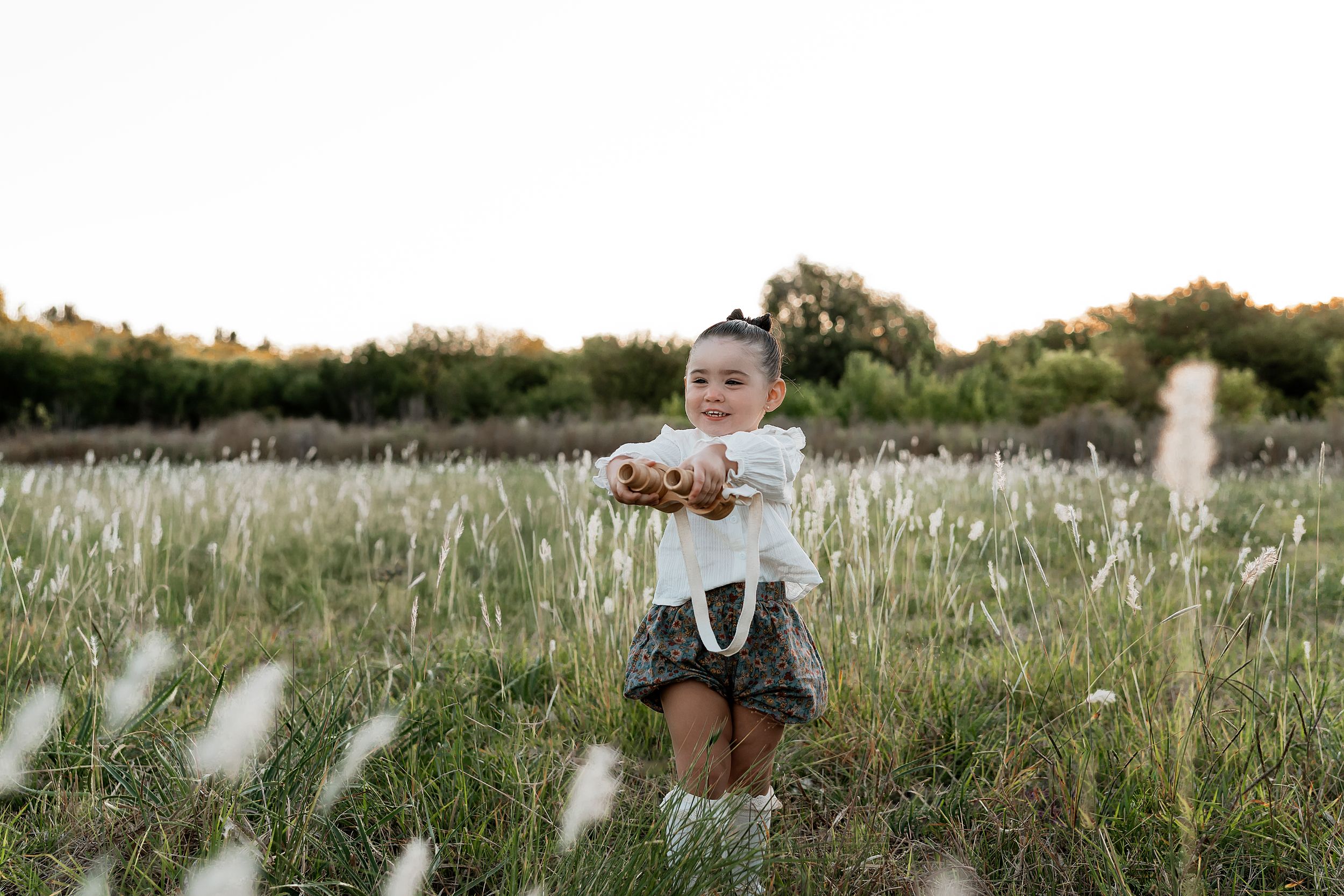 Toddler girl in white top and plaid skirt holding a toy in a scenic grass field at golden hour.