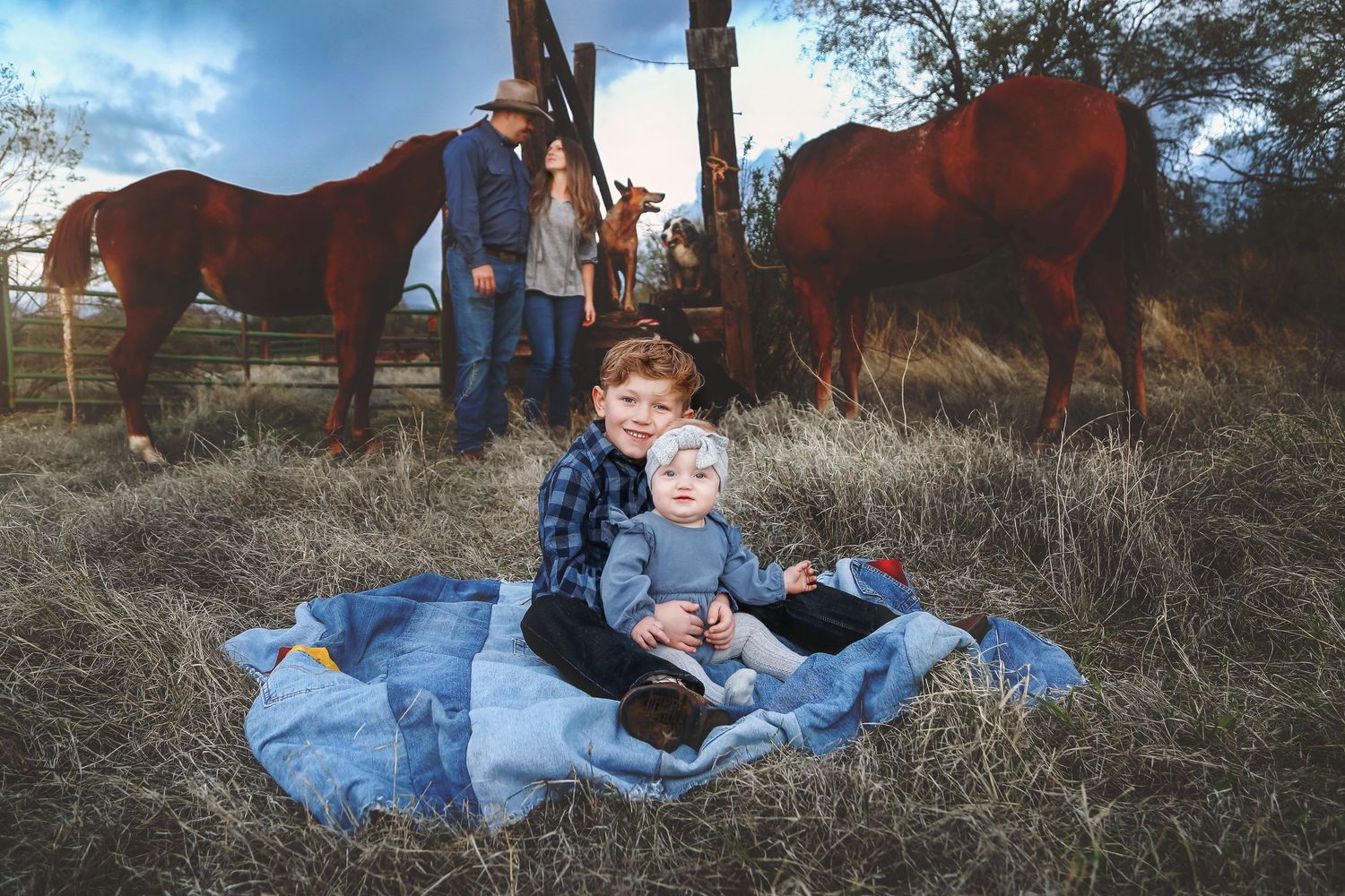 Two young children sit on a blue blanket in a grassy field while two chestnut horses graze and a man and woman stand by a wooden fence.