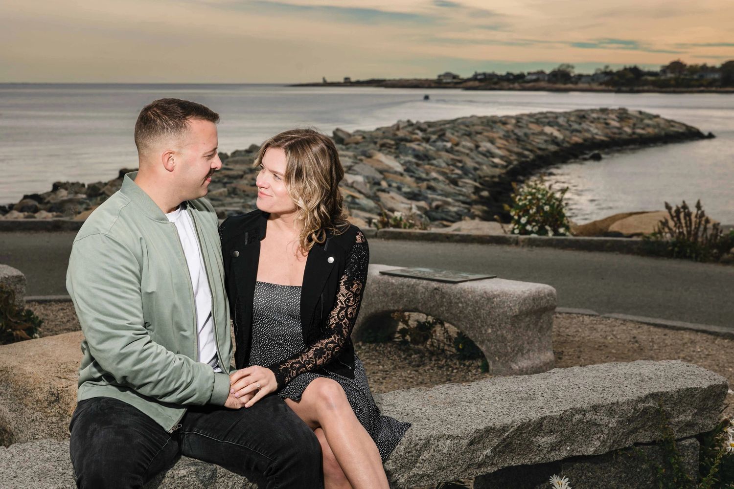 Couple sitting together on a weathered bench, gazing toward the horizon over water. Engagement at Rockport Harbor, Rockport, MA