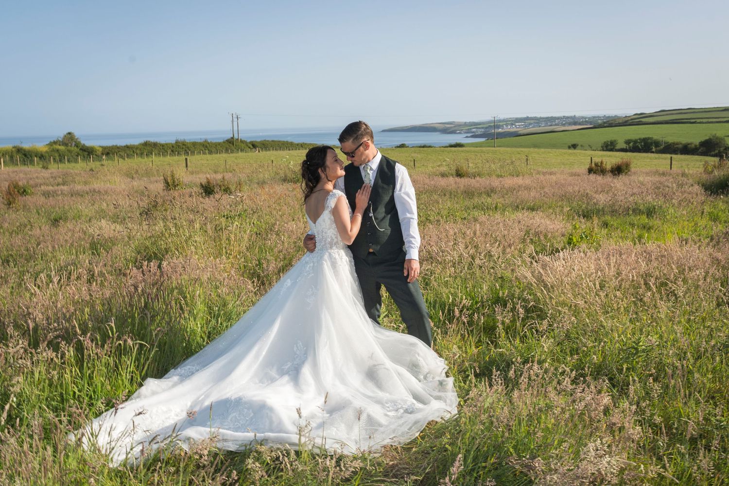 Bride in a white gown and groom in a gray vest pose together beside a still pond with reflections, surrounded by lush ferns and palms.