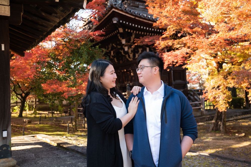Smiling couple embracing in front of a traditional Japanese temple surrounded by vibrant red autumn maple leaves.