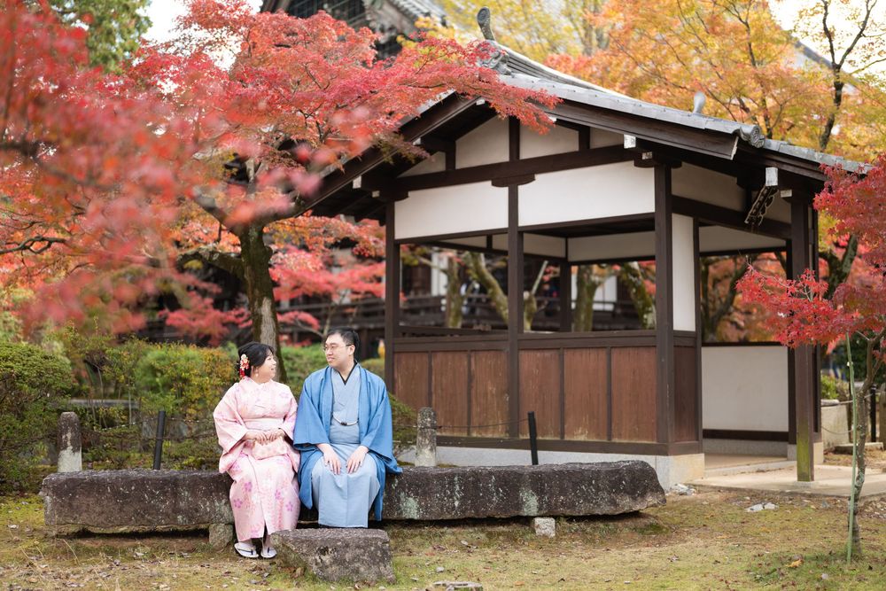 Two people in traditional Japanese kimono sit on a stone bench beside a wooden pavilion surrounded by vibrant red autumn maple leaves.