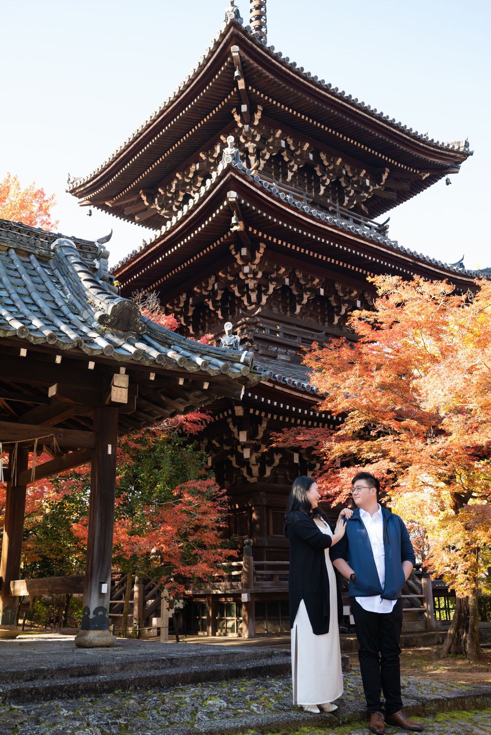 A couple poses in front of a traditional multi-tiered Japanese pagoda surrounded by vibrant red autumn maple leaves.
