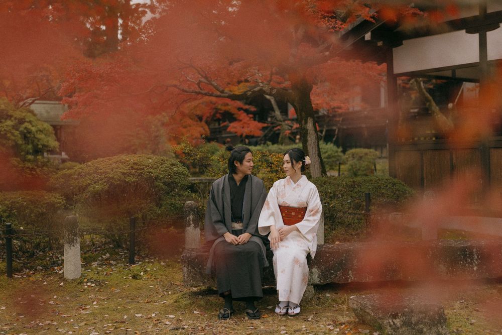 Couple in traditional Japanese kimono sitting together on a stone bench beneath vibrant red autumn maple leaves in a serene garden.