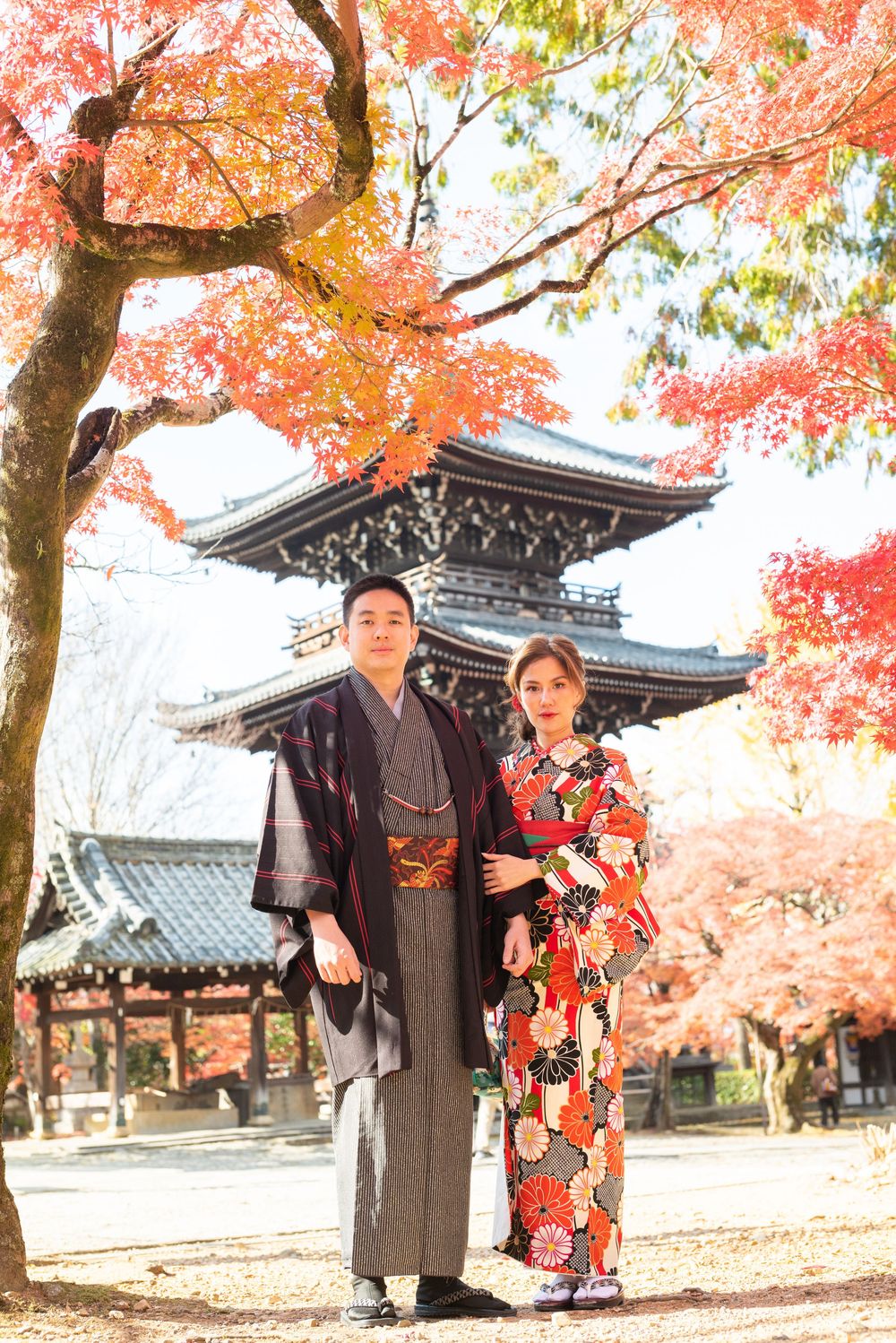 Couple in traditional Japanese kimono stand beneath vibrant red autumn maple leaves with a historic pagoda in the background.