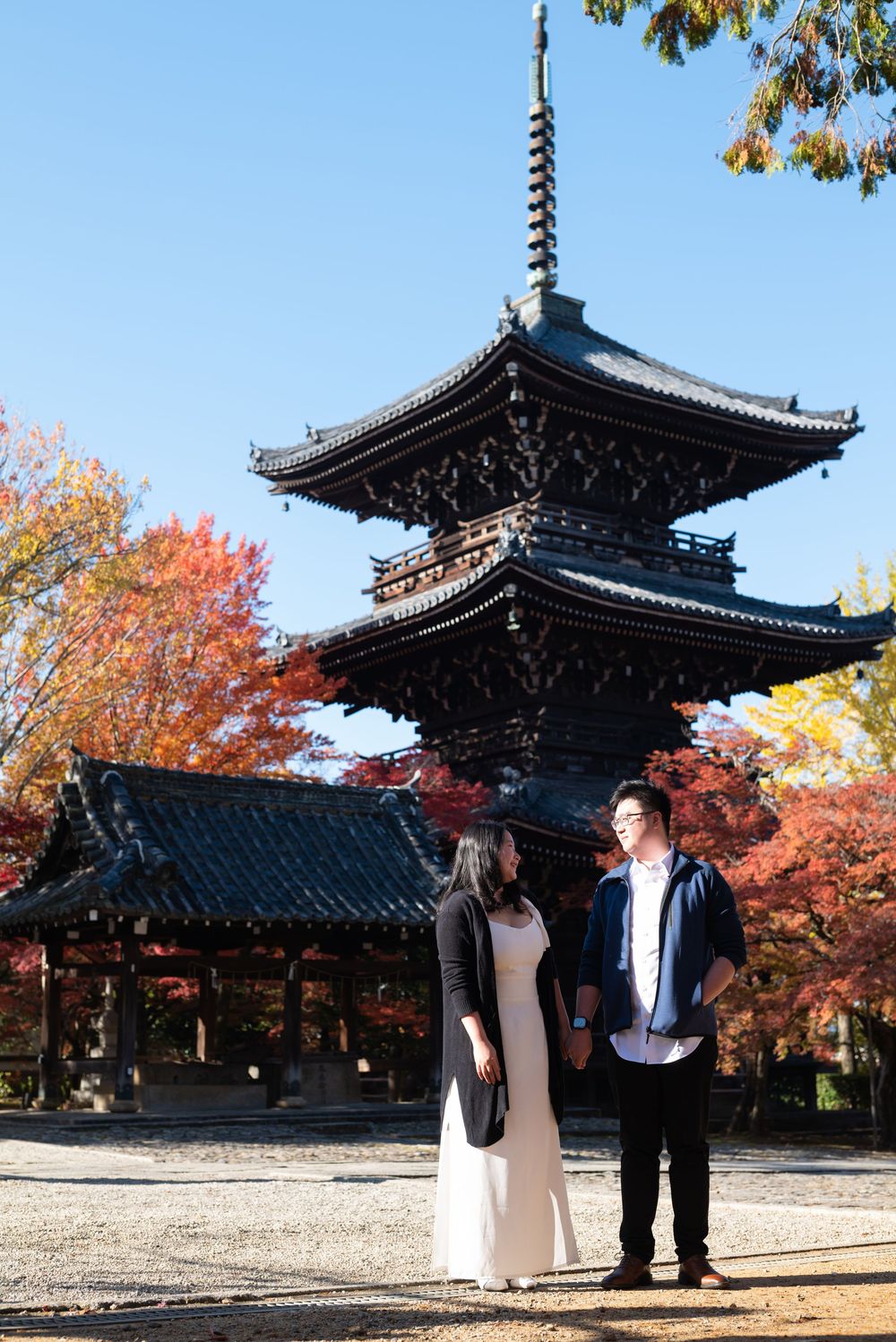 Couple stands hand in hand before a multi-tiered Japanese pagoda surrounded by vibrant red autumn maple leaves.