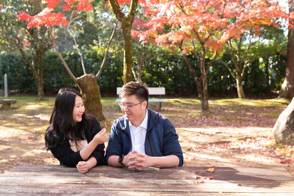 Smiling couple sitting at a wooden picnic table in a park surrounded by vibrant red autumn maple leaves.
