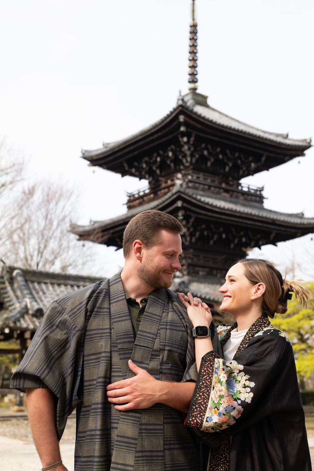 Smiling couple in traditional Japanese kimono-style outfits embrace in front of a historic multi-tiered pagoda on a bright day in Kyoto.