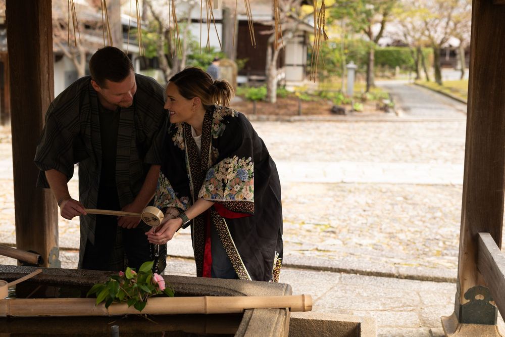 Couple in traditional Japanese kimono purify their hands with bamboo ladles at a temple water basin in a peaceful courtyard.