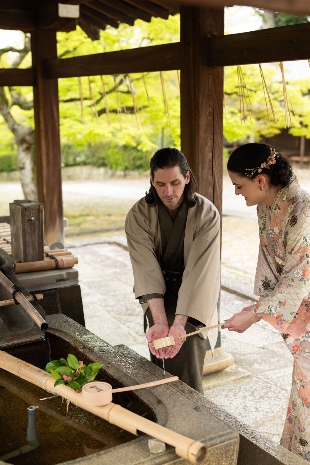Man in a kimono-style robe and woman in a floral kimono purify their hands with bamboo ladles at a stone water basin in a Japanese shrine.