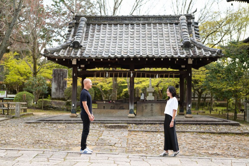 A man and a woman stand facing each other in front of a traditional Japanese temple pavilion with a tiled roof surrounded by autumn trees.