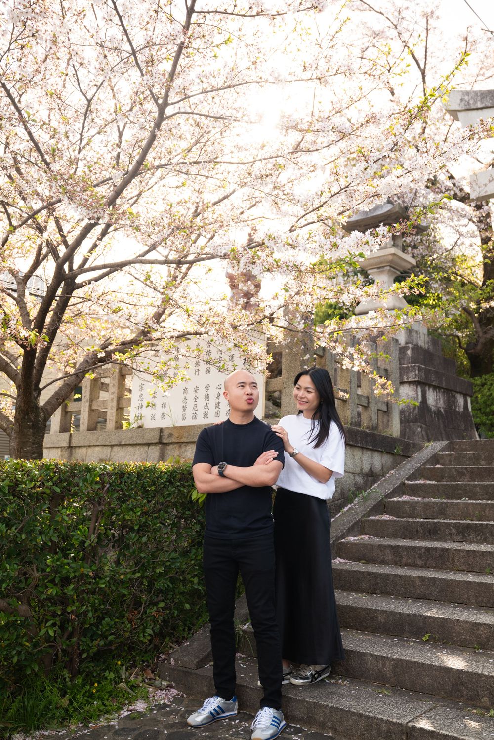 Couple posing on stone steps beneath blooming cherry blossom trees near a traditional Japanese shrine wall.