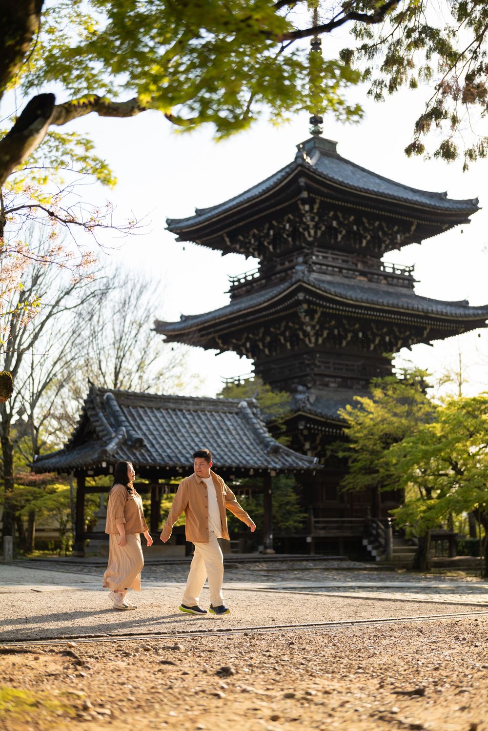 Couple in beige outfits stroll hand in hand past a traditional Japanese temple with a two-story pagoda surrounded by spring trees.