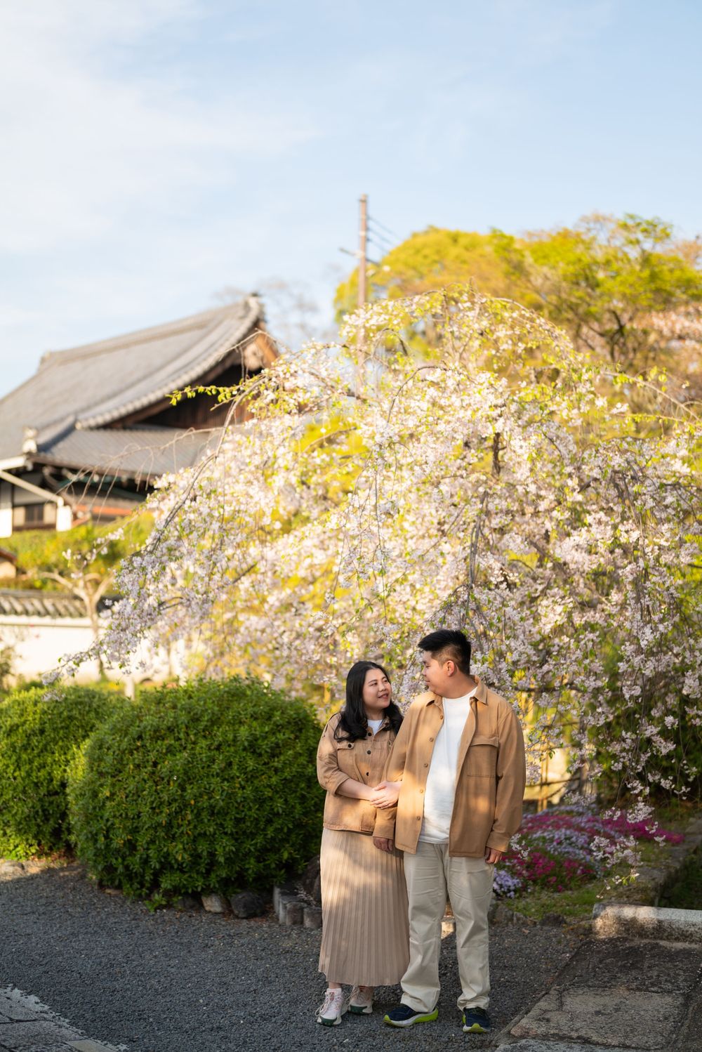 Smiling couple in matching tan jackets stand together beneath a blooming cherry blossom tree near a traditional Japanese temple.
