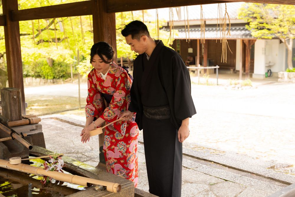 Couple in traditional Japanese kimono and black haori standing beside a stone water basin at a serene temple garden.