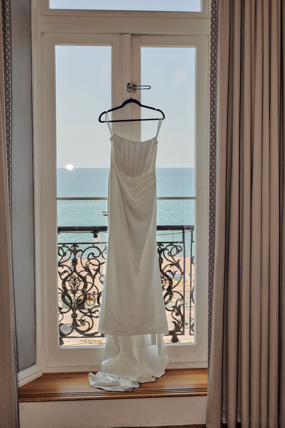 White strapless wedding dress hanging in window overlooking the sea on a bright day.