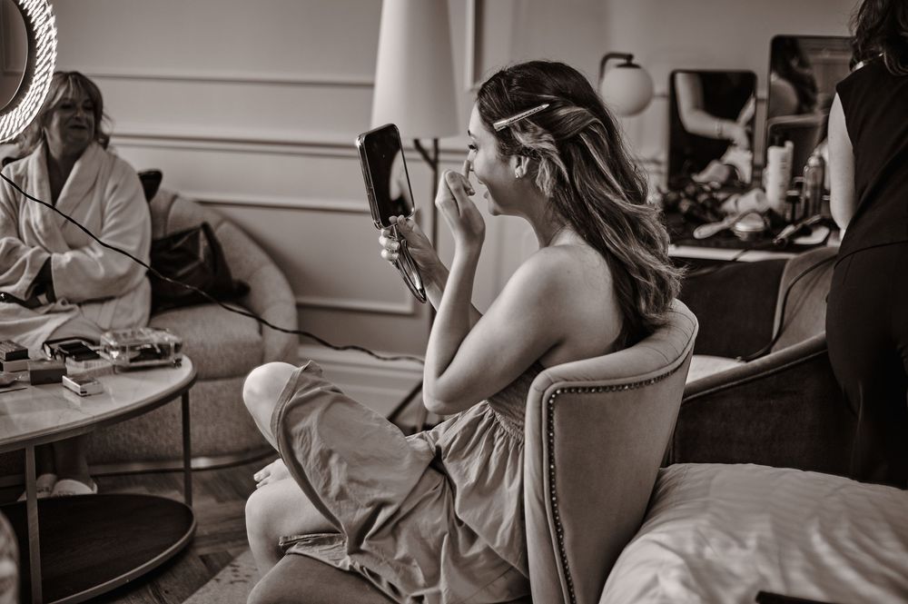 Black and white photo of a bride applying makeup while seated at a vanity mirror.