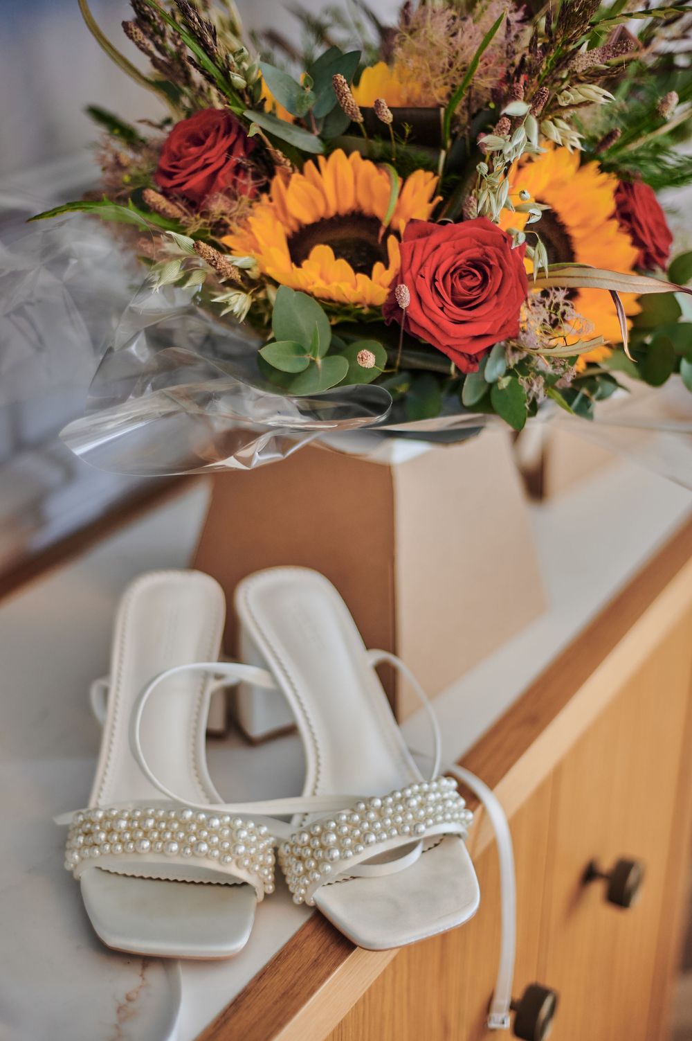 Pearl-embellished bridal sandals on a dresser beside a sunflower and red rose wedding bouquet.