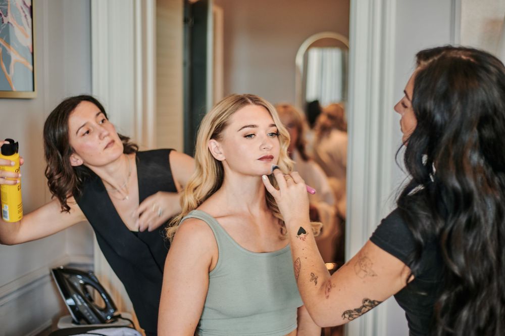 Bride gets makeup applied by artist while hairstylist works in bright dressing room.