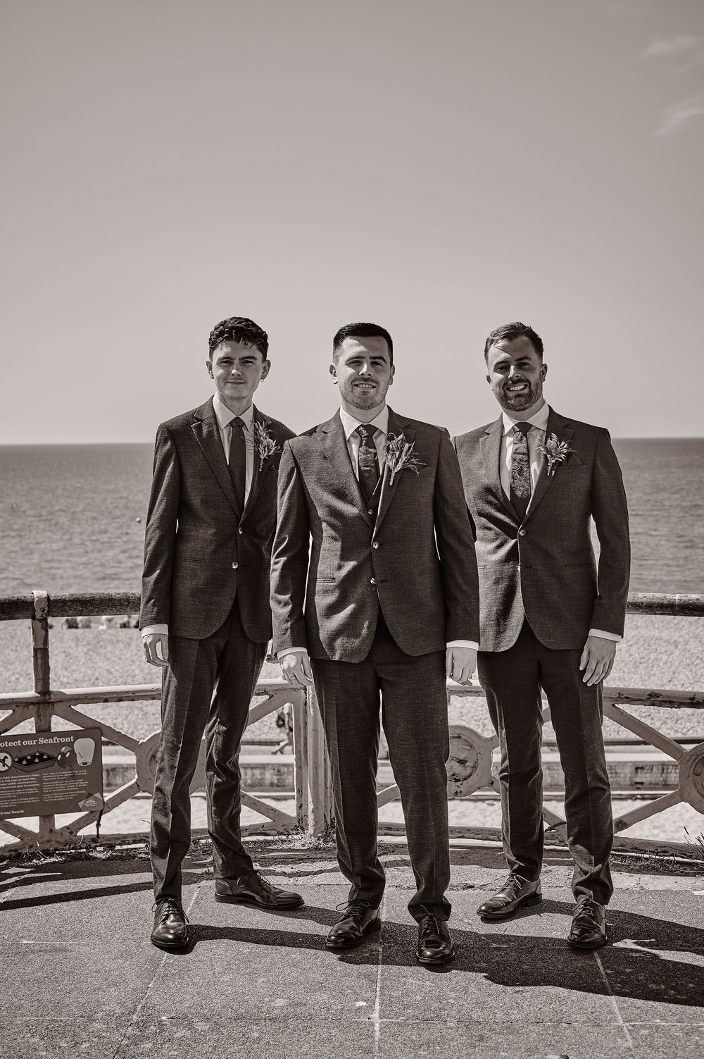 Groom and two groomsmen in suits posing by the seaside in a black and white photo.